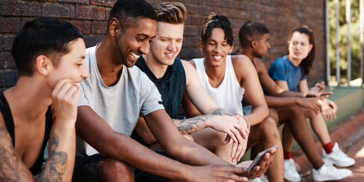 Symbolbild, Gruppe an jungen Sportlerinnen und Sportlern sitzt lachend zusammen und schaut auf ein Smartphone