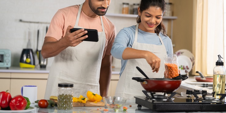 Symbolbild, indisches Pärchen kocht gemeinsam in der Küche. Der Mann schaut dabei auf sein Smartphone.