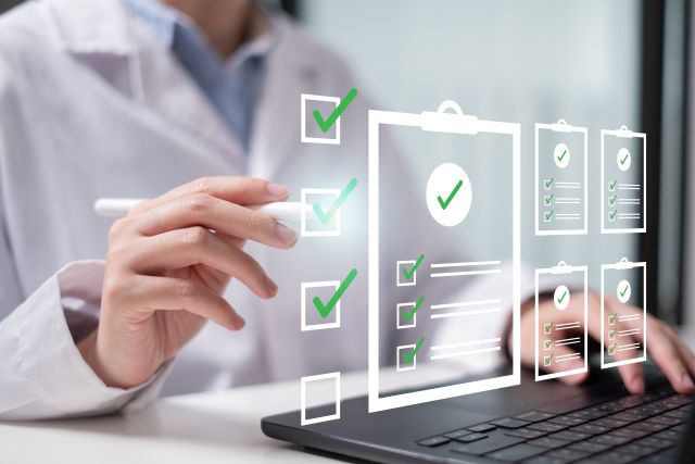 Person im weißen Kittel zeigt auf digitale Checkliste mit grünen Häkchen vor einem Laptop