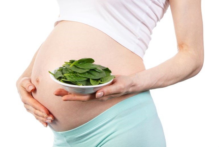 Schwangere Frau in weißem Top und türkiser Hose hält Schüssel mit frischem Spinat vor Babybauch.