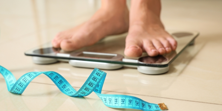 Symbolbild, Füße eines Menschen stehen auf einer Personenwaage und davor liegt ein ausgerolltes Maßband