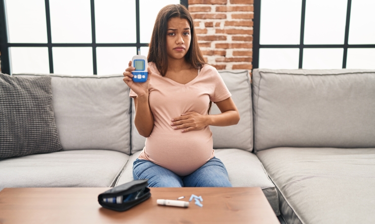 Symbolbild, Schwangere sitzt mit Blutgkucometer in der Hand auf dem Sofa und schaut skeptisch die die Kamera