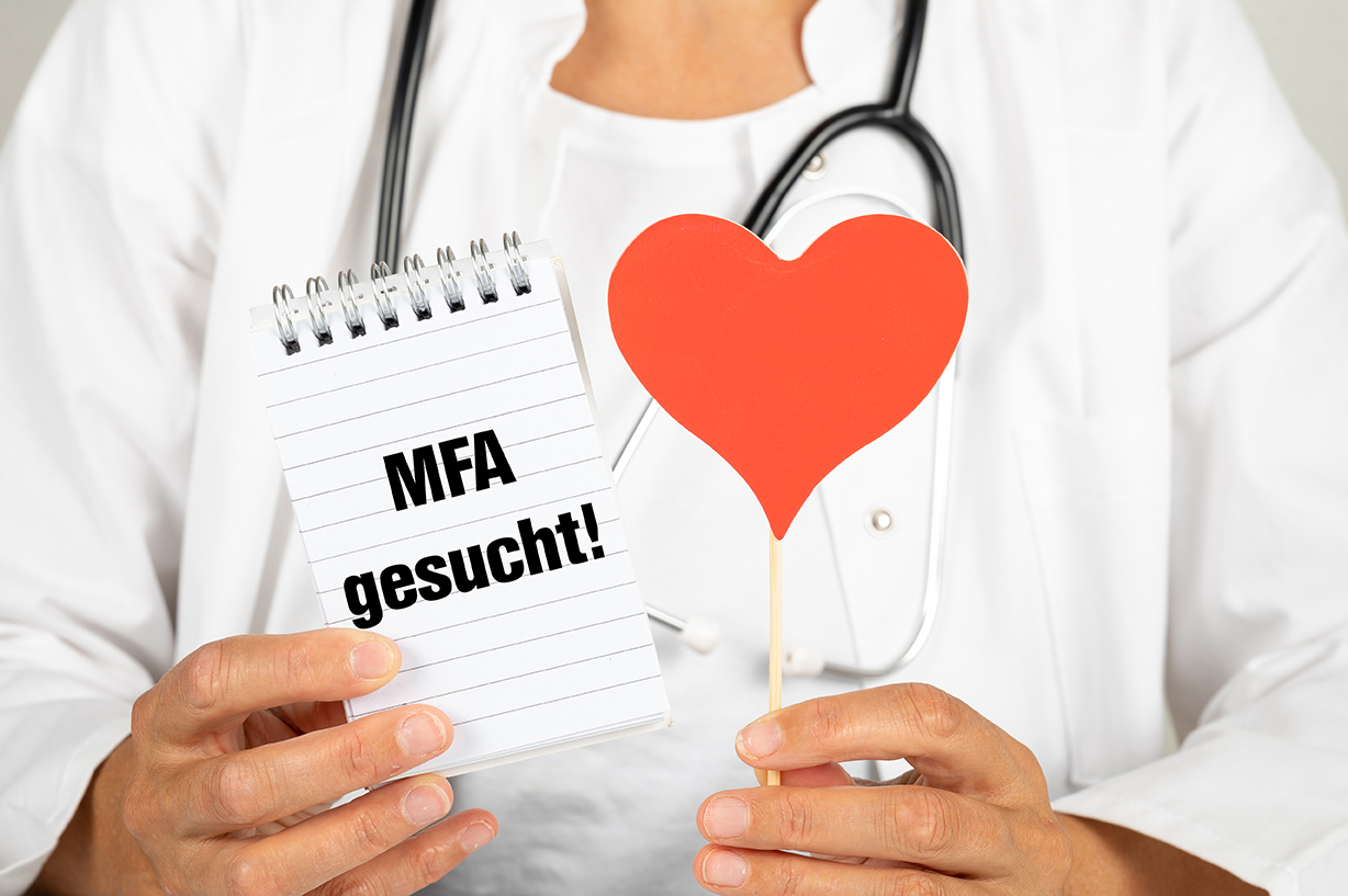 Eine Person im Arztkittel hält einen Notizblock mit der Aufschrift "MFA gesucht!" und ein rotes Herz.