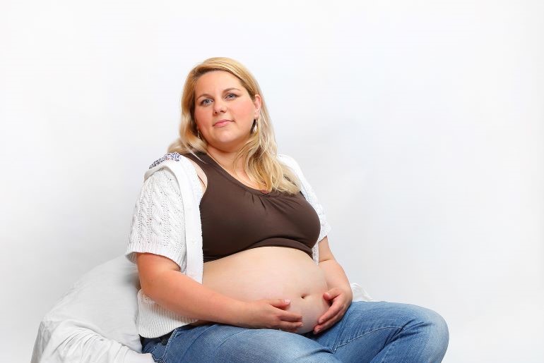 Übergewichtige Schwangere lächelt trotz Stoffwechselrisiko, sitzt am Boden und hält ihren Babybauch.