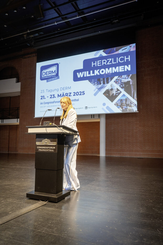 Rednerin am Pult während der 23. Tagung DERM 2025 im CongressForum Frankenthal mit einer großen Leinwand im Hintergrund.