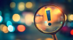 Lupe mit einem Ausrufezeichen im Fokus, vor einem unscharfen, farbenfrohen Hintergrund.