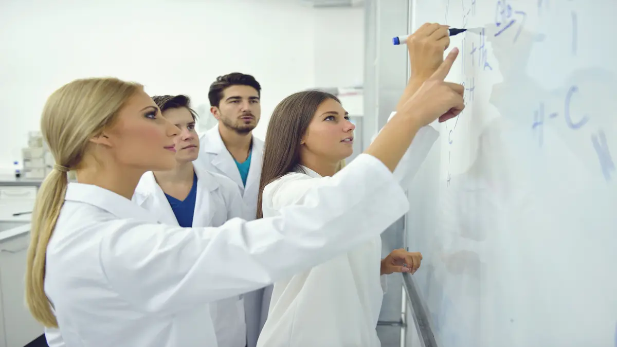 Mehrere Studierende in weißen Laborkitteln stehen in einem modernen Labor und arbeiten zusammen an einem Whiteboard. Eine Person schreibt mit einem Marker Formeln an die Tafel, während die anderen aufmerksam zusehen und mitdiskutieren. Die Umgebung wirkt hell und wissenschaftlich.