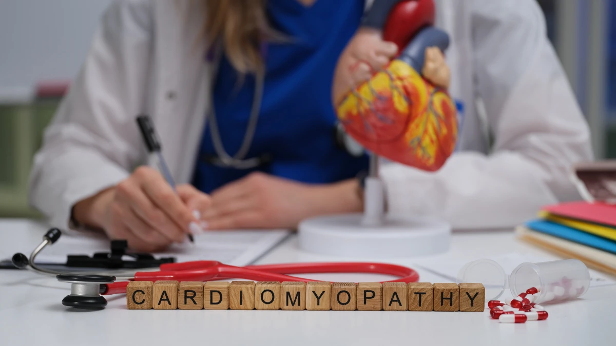 Eine Ärztin macht sich Notizen am Schreibtisch. Vor ihr stehen ein anatomisches Herz-Modell und das Wort "Cardiomyopathy" in Holzbuchstaben.