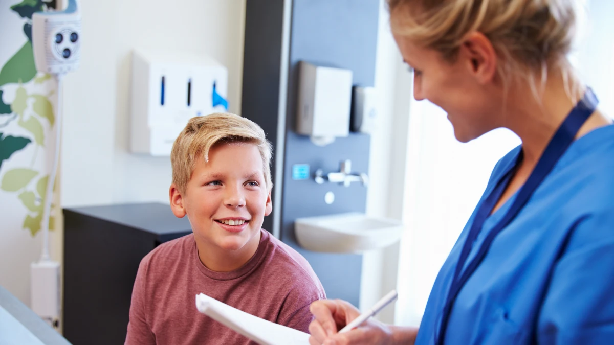 Krankenpflegerin unterhält sich mit einem Jungen und macht sich dabei Notizen.