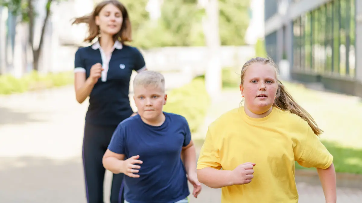 ein Junge und Mädchen mit Übergewicht joggen mit einer Trainerin im Hintergrund